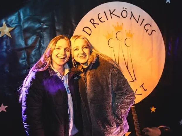 Zwei l&auml;chelnde Frauen stehen dicht nebeneinander und blicken in die Kamera. Sie tragen warme Jacken und stehen vor einer dekorierten B&uuml;hne mit Sternen, einem gro&szlig;en Mondmotiv mit der Aufschrift &bdquo;Dreik&ouml;nigs&ldquo; und einer im Hintergrund sichtbaren Gitarre